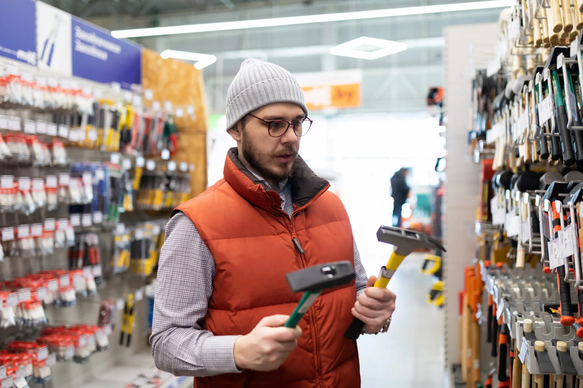 A man stands in the isle of a hardware store and chooses between two hammers